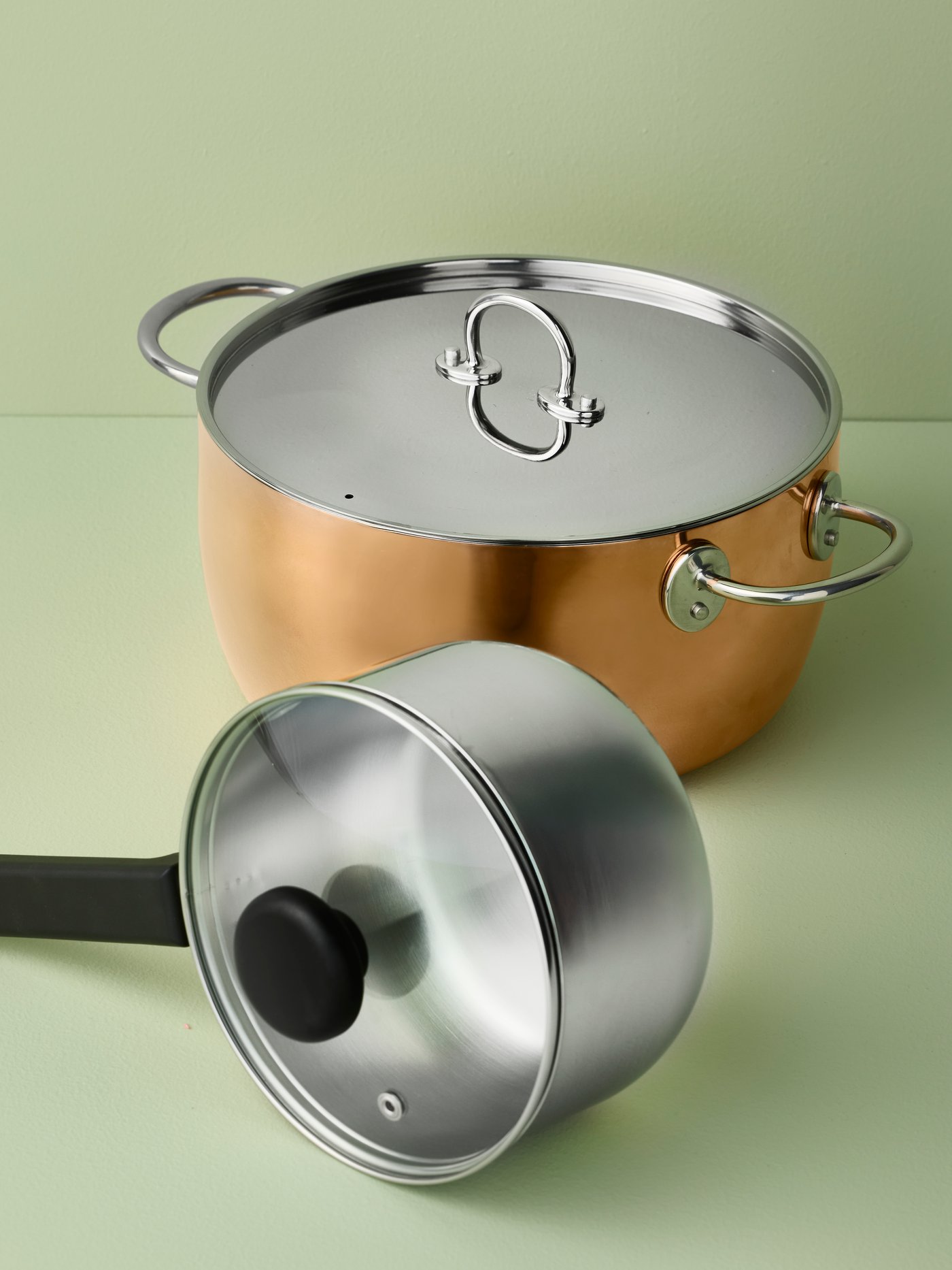 A MIDDAGSMAT saucepan with lid and a FINMAT pot with lid in copper and stainless steel are placed on a light green surface.