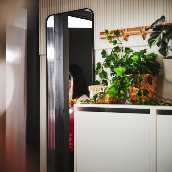 Miroir LINDBYN noir près d’une porte d’entrée ouverte, et deux armoires à chaussures BISSA sur lesquelles sont disposées des plantes artificielles en pot FEJKA.