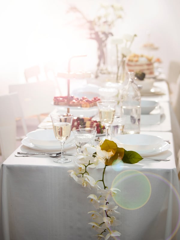 Softly lit dining table with white tablecloth set with white OFTAST dishes for multiple guests. Flowers spill over the side of the table and placed in vases behind.