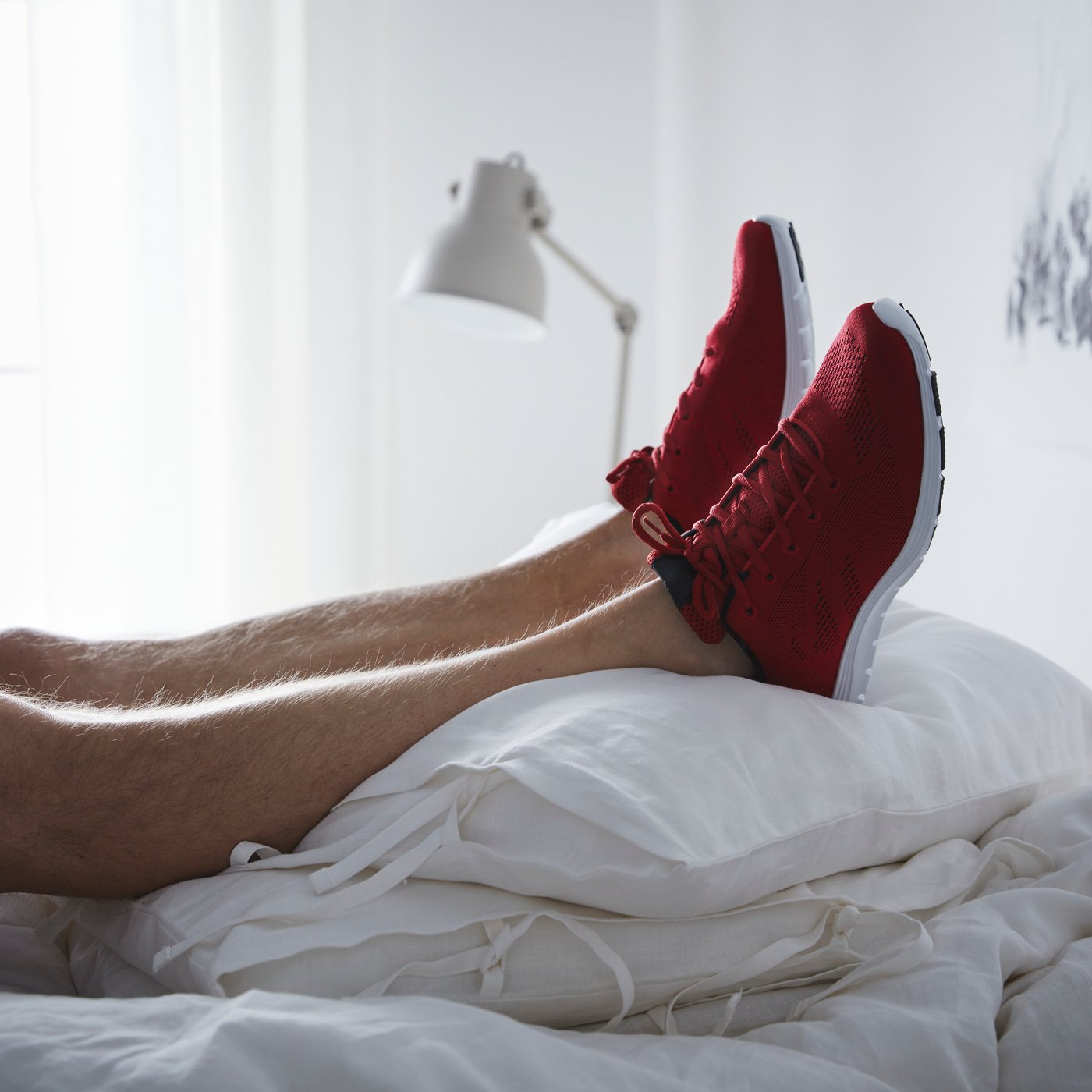 Un par de pies con zapatillas deportivas de color rojo brillante se encuentran encima de una pila de almohadas blancas y frescas, con una lámpara al fondo.