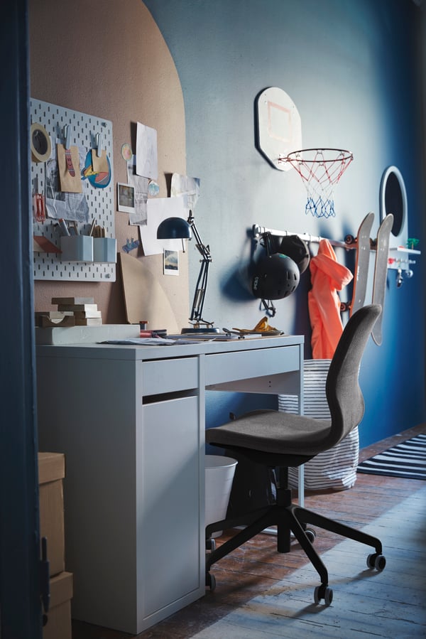 Uma secretária alta em madeira com três níveis, um portátil e um candeeiro no tampo e diversos artigos em baixo, em frente a uma parede azul.