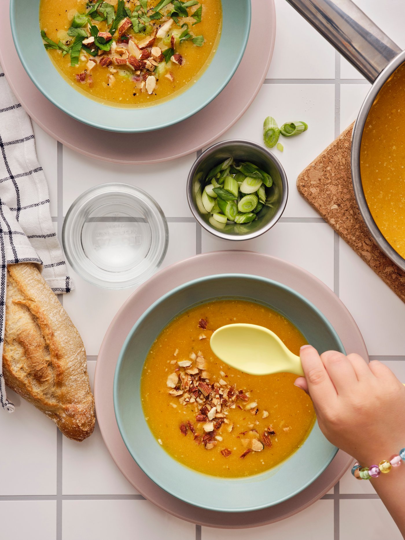 A hand holding a spoon scoops up pumpkin soup in a light turquoise FÄRGKLAR deep plate, placed on a tiled table.