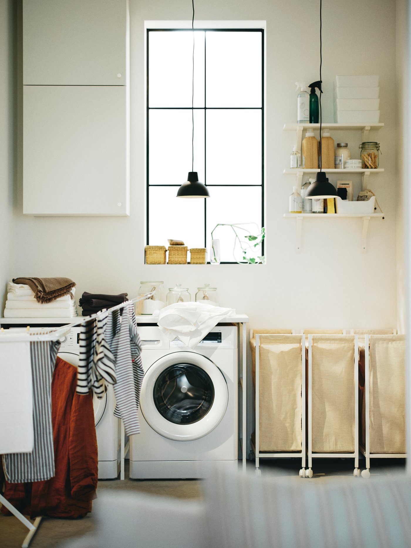 큰 창문이 있는 세탁실에 드럼세탁기와 오른쪽으로 엔헤트 빨래바구니 3개가 놓여있는 모습.