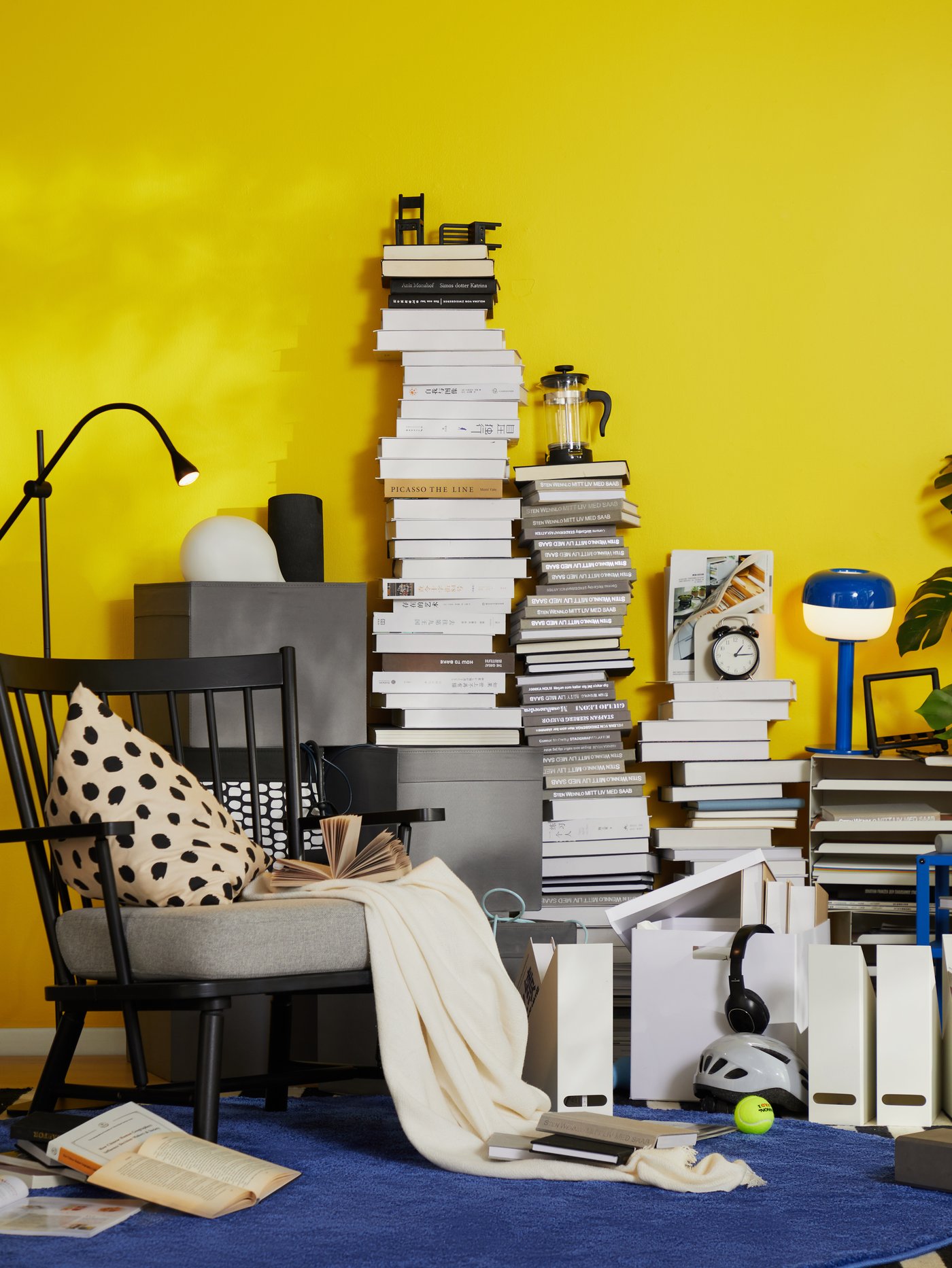 Dnevna soba, zapolnjena z neurejenimi kupi knjig in revij pred rumeno steno, ob kateri je črn fotelj.