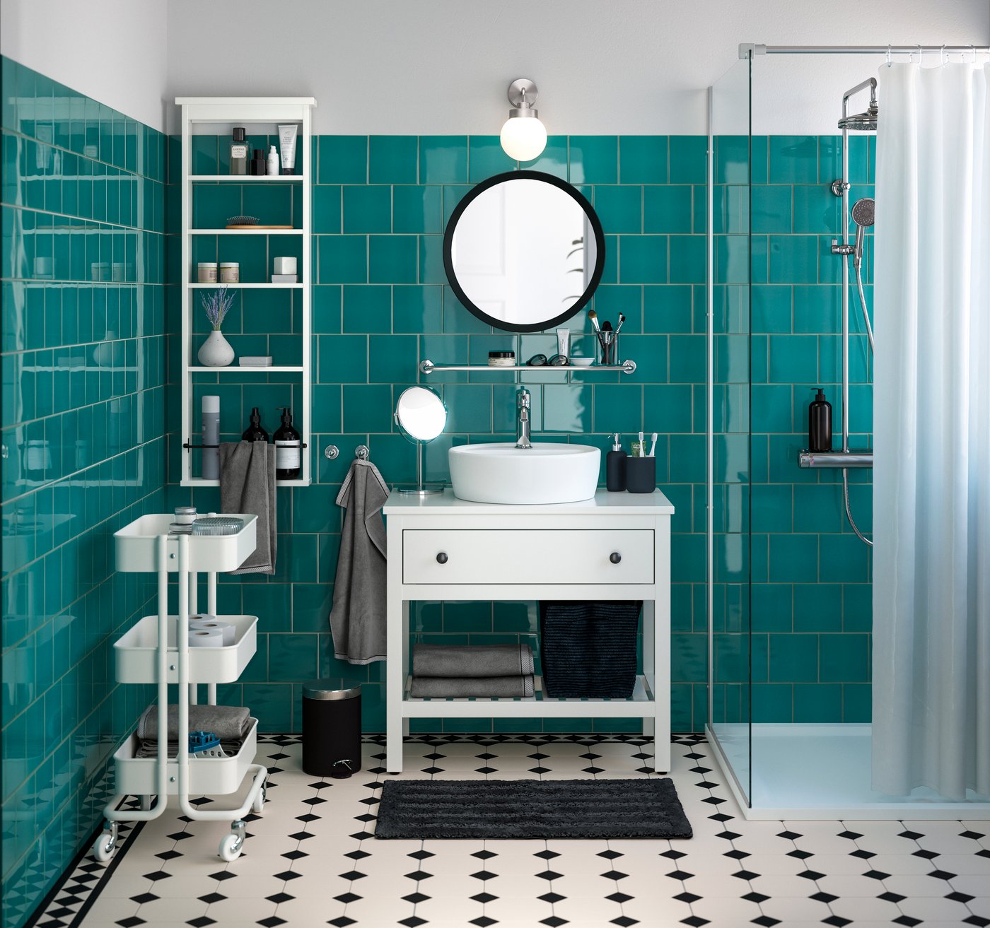 Un bagno HEMNES bianco visto frontalmente completo di lavabo in ceramica rotondo bianco, mobile per lavabo con un cassetto, specchio rotondo e pensile a giorno. Di fianco un box doccia e un carrello portatutto bianco.