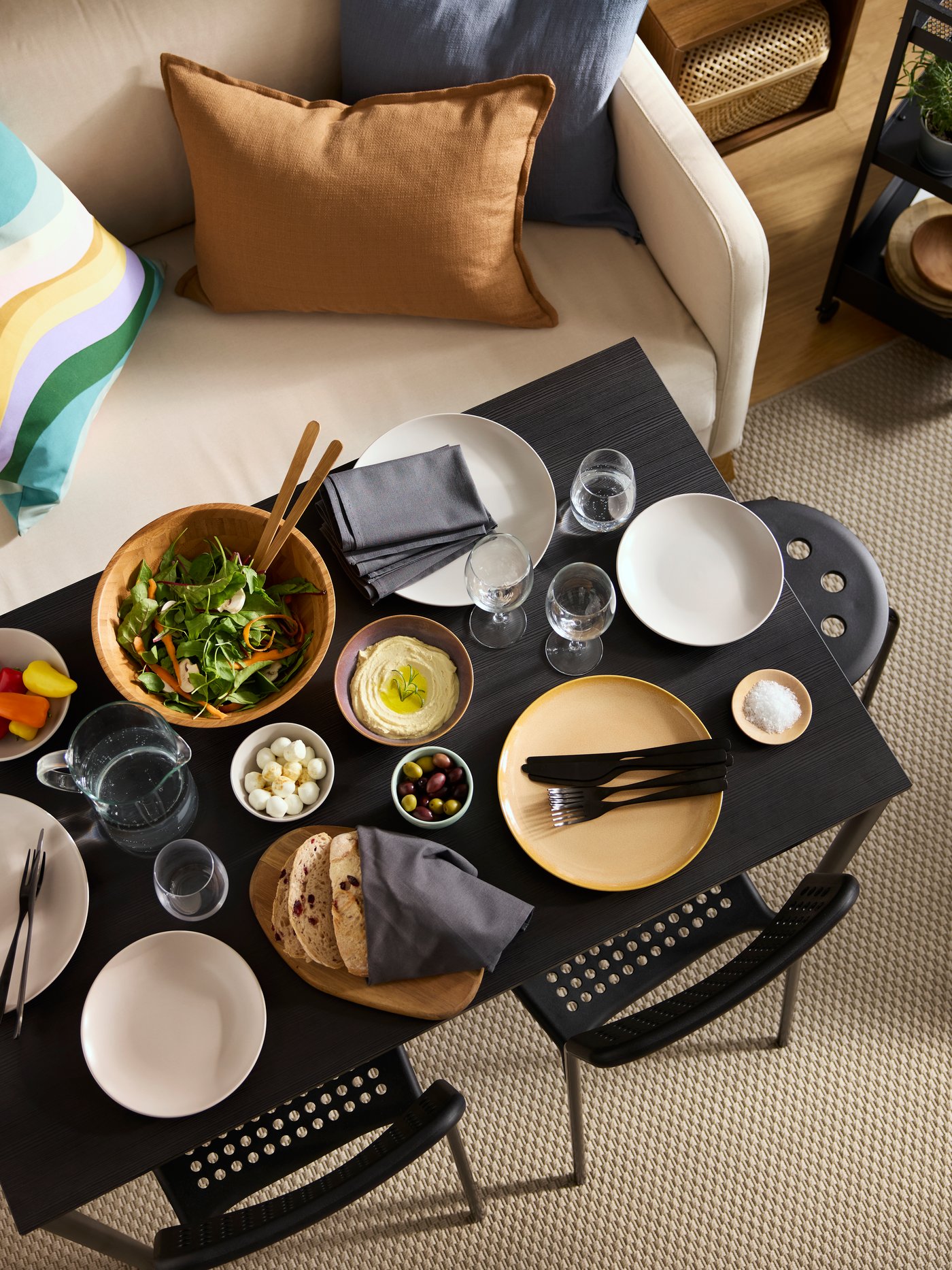 Un repas coloré servi avec des assiettes FÄRGKLAR sur une table noire SANDSBERG flanquée de chaises ADDE, d'un tabouret MARIUS et d'un canapé.