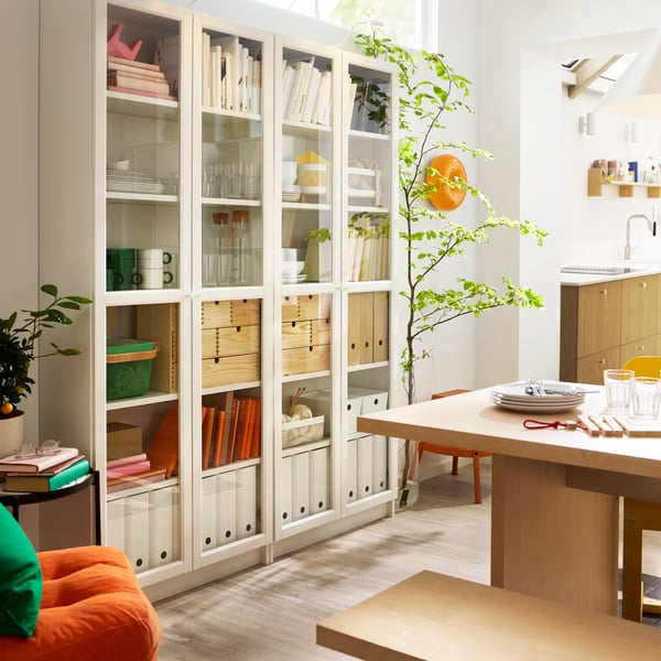 White BILLY bookcases with glass doors, placed next to a table, have binders, boxes, glasses and mugs placed on the shelves.