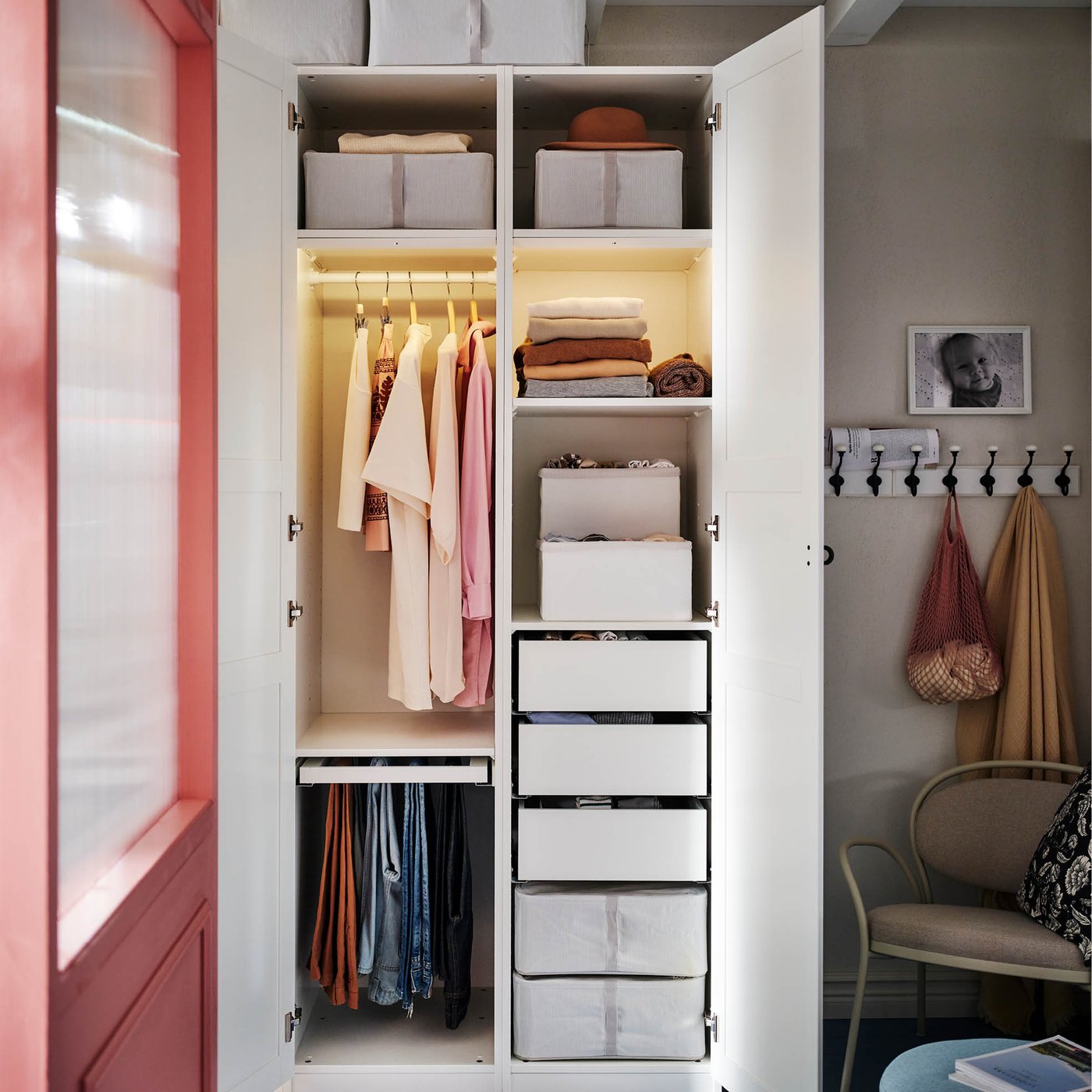 An open white IKEA wardrobe, organized with hanging clothes, folded garments, storage boxes, and drawers, stands beside a pink door and a wall with a coat rack and a chair.