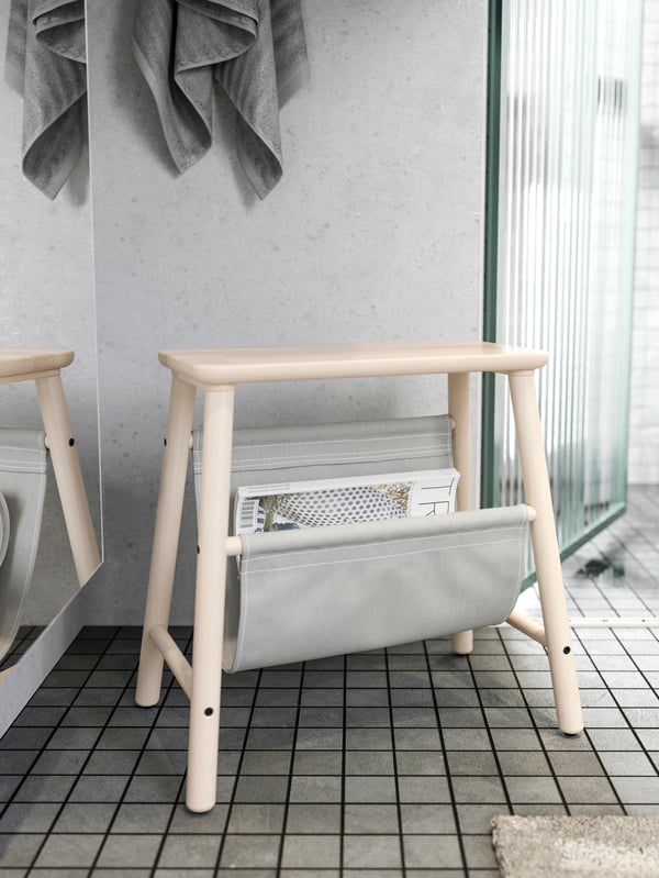 A VILTO storage stool in birch with a pocket containing a magazine, on a tiled bathroom floor next to a shower door.
