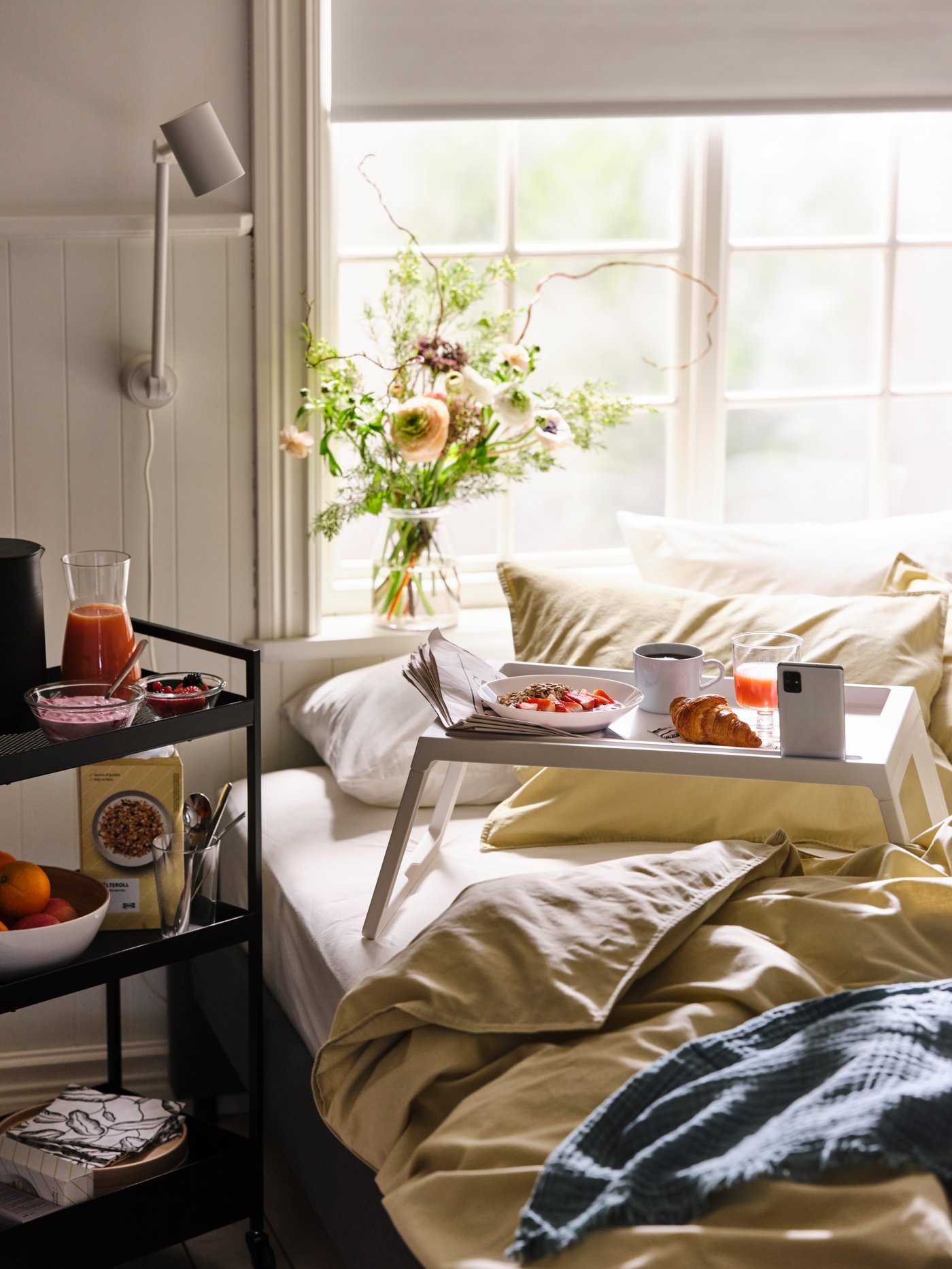 Carrito NISSAFORS negro con artículos de desayuno, junto a una bandeja de desayuno llena sobre una cama hecha con ropa de cama ÄNGSLILJA .