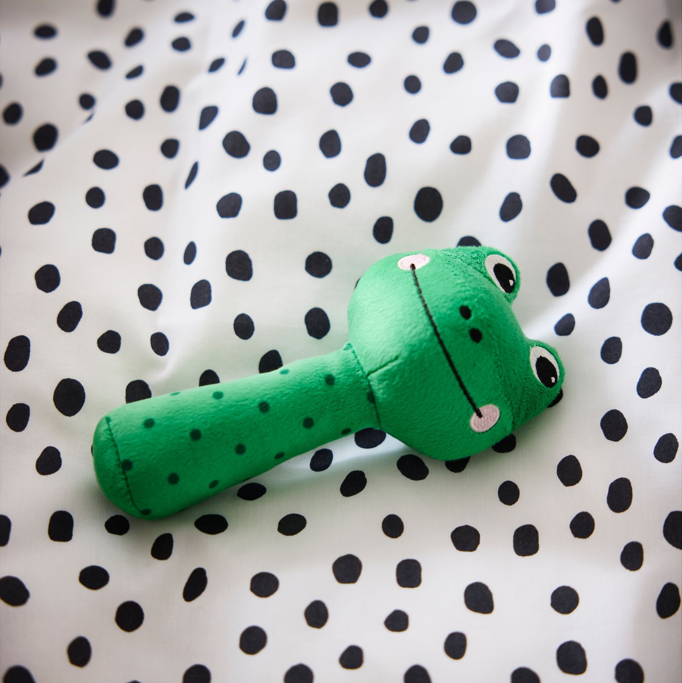 A close-up of SANDBI rattle in the shape of a green frog, laying on a white duvet cover with small black dots.