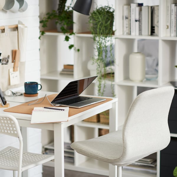 A white swivel chair stands at a white MELLTORP table with a laptop on it near a white KALLAX shelving unit.