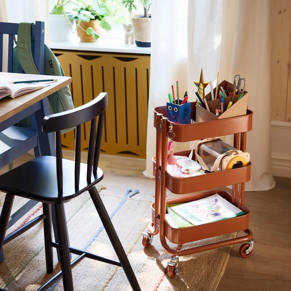 Un chariot RÅSKOG marron-rouge contenant des crayons, des livres et d'autres objets, se tient à côté d'une chaise junior AGAM sur un tapis RAKLEV.