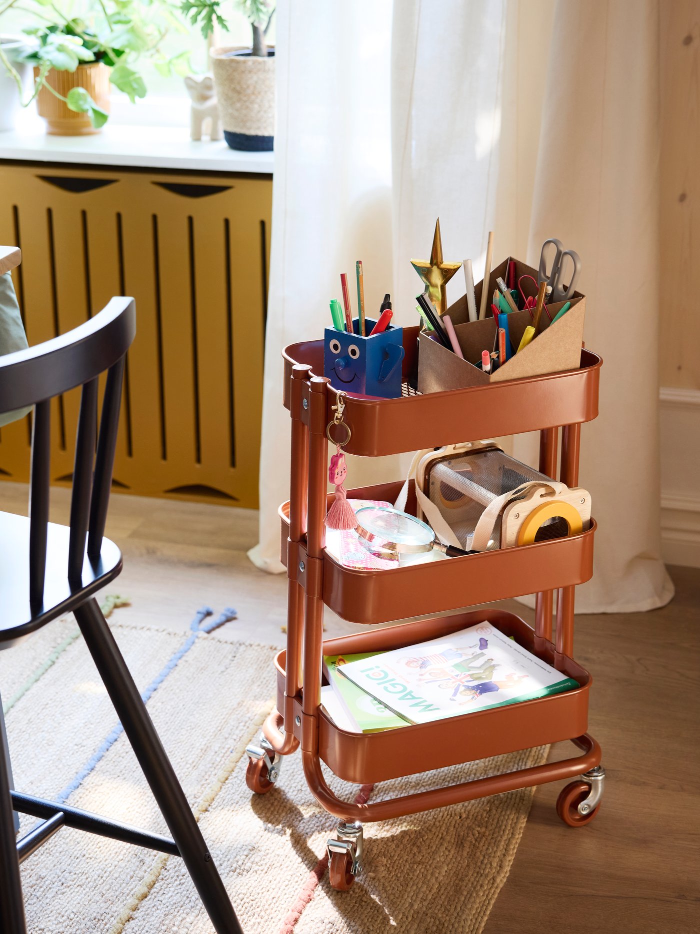 Ein rotbrauner RÅSKOG-Rollwagen, in dem Bleistifte, Bücher und andere Dinge liegen, steht neben einem AGAM-Junior-Stuhl auf einem RAKLEV-Teppich.