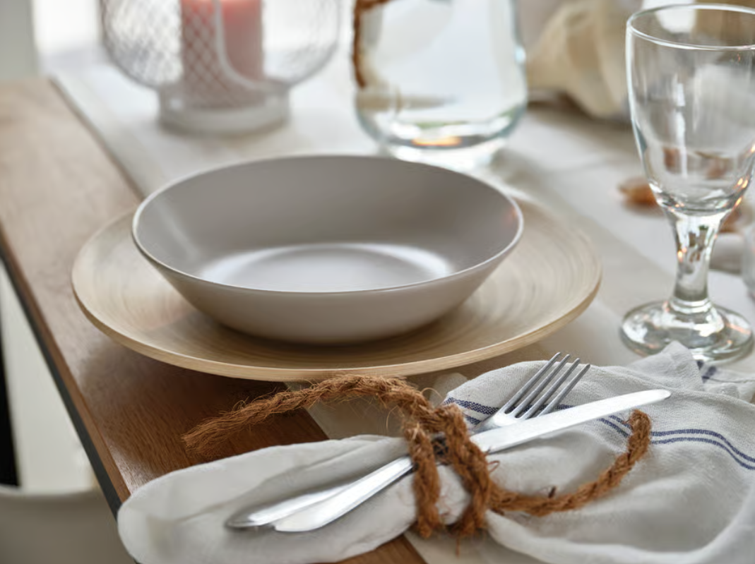 Tischgedeck mit heller Schale auf Holzuntersetzer, Weinglas und mit Kordel gebundenem Besteck auf hell gestreiftem Läufer.
