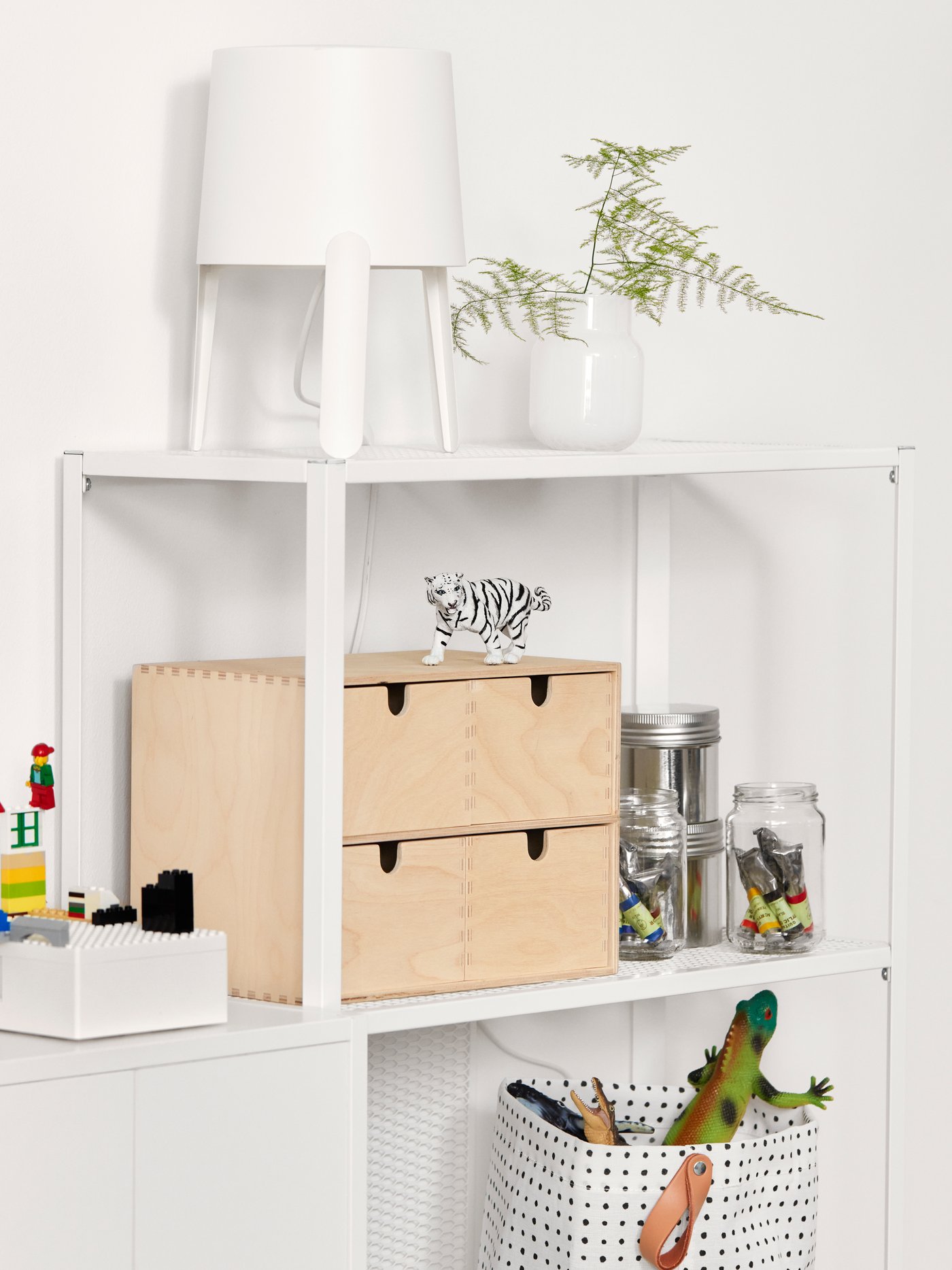 Una estantería blanca, una cesta de almacenaje en blanco/negro con dinosaurios de juguete, botes transparentes con tubos de pintura y una lámpara de mesa blanca.