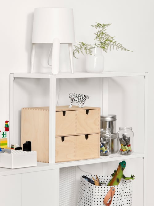 A white shelving unit, a white/black storage basket with dinosaur toys, clear jars with paint tubes, a white table lamp.