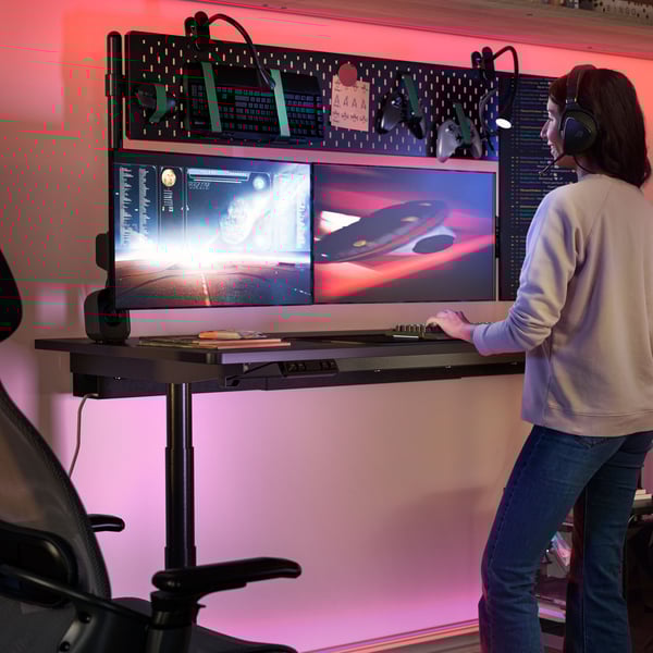 A person is standing at a MÅLOMRÅDE sit/stand gaming desk, playing an online game. Pink light glows behind the desk.