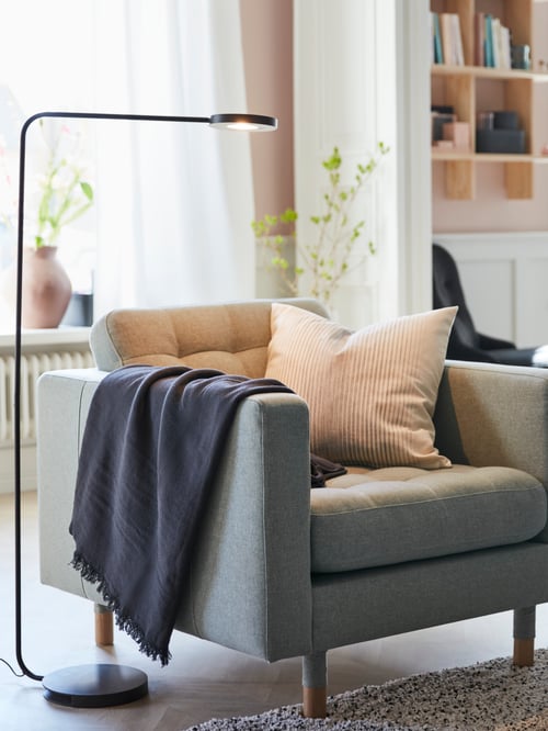 Living room with armchair with armrests, LED floor lamp, cushion cover, throw, shelving unit, plants in pots, white curtains.