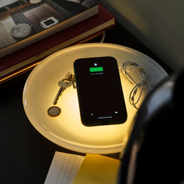 A lit-up VÄSTMÄRKE wireless charger with lighting charges a mobile phone. Keys lie on the bowl of the charger.