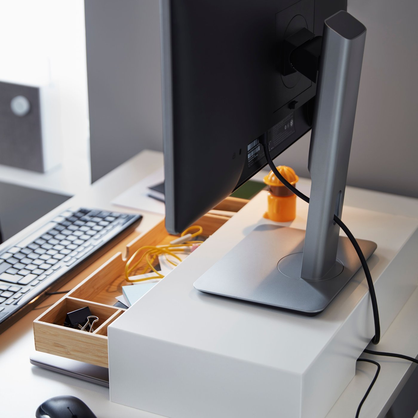Eine weiße ELLOVEN Monitorerhöhung mit Schublade steht auf einem Schreibtisch, auf dem sich auch ein Monitor befindet. Die Schublade steht offen.