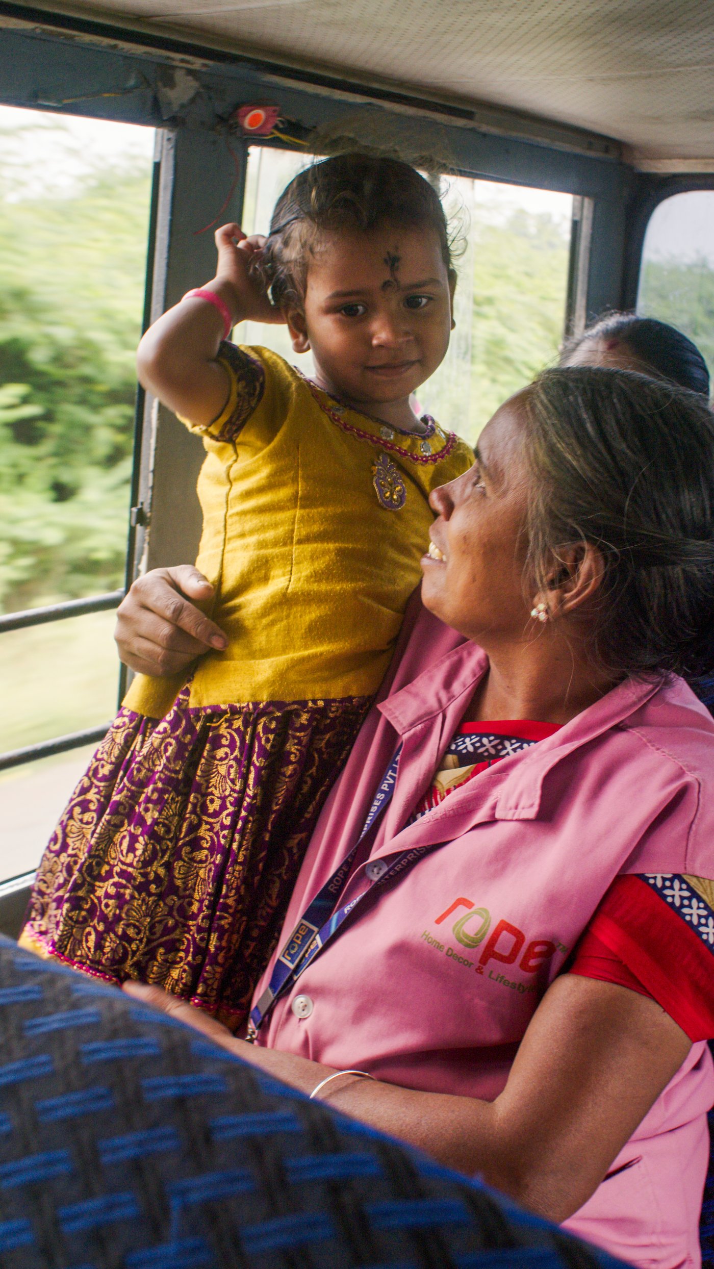 A person in pink clothes stands smiling holding a small child in her arms inside a bus