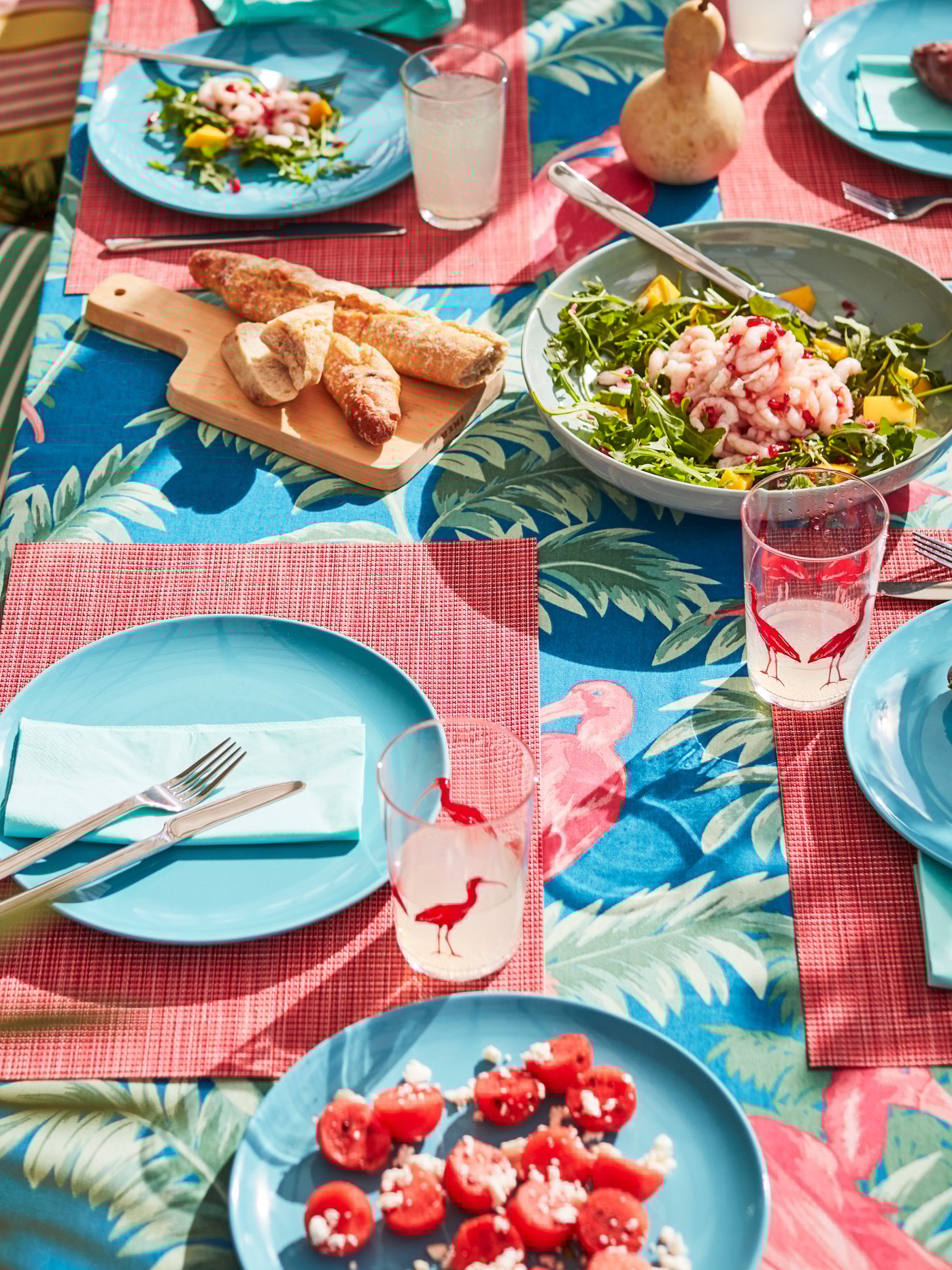 A sunny, summery table set with turquoise plates, light red place mats and a multi-colored tablecloth with flamingos.
