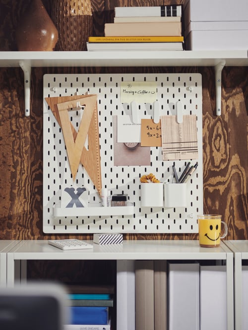 Close-up on workspace wall with shelves and white wooden pegboard mounted on the wall holding accessories and notes.