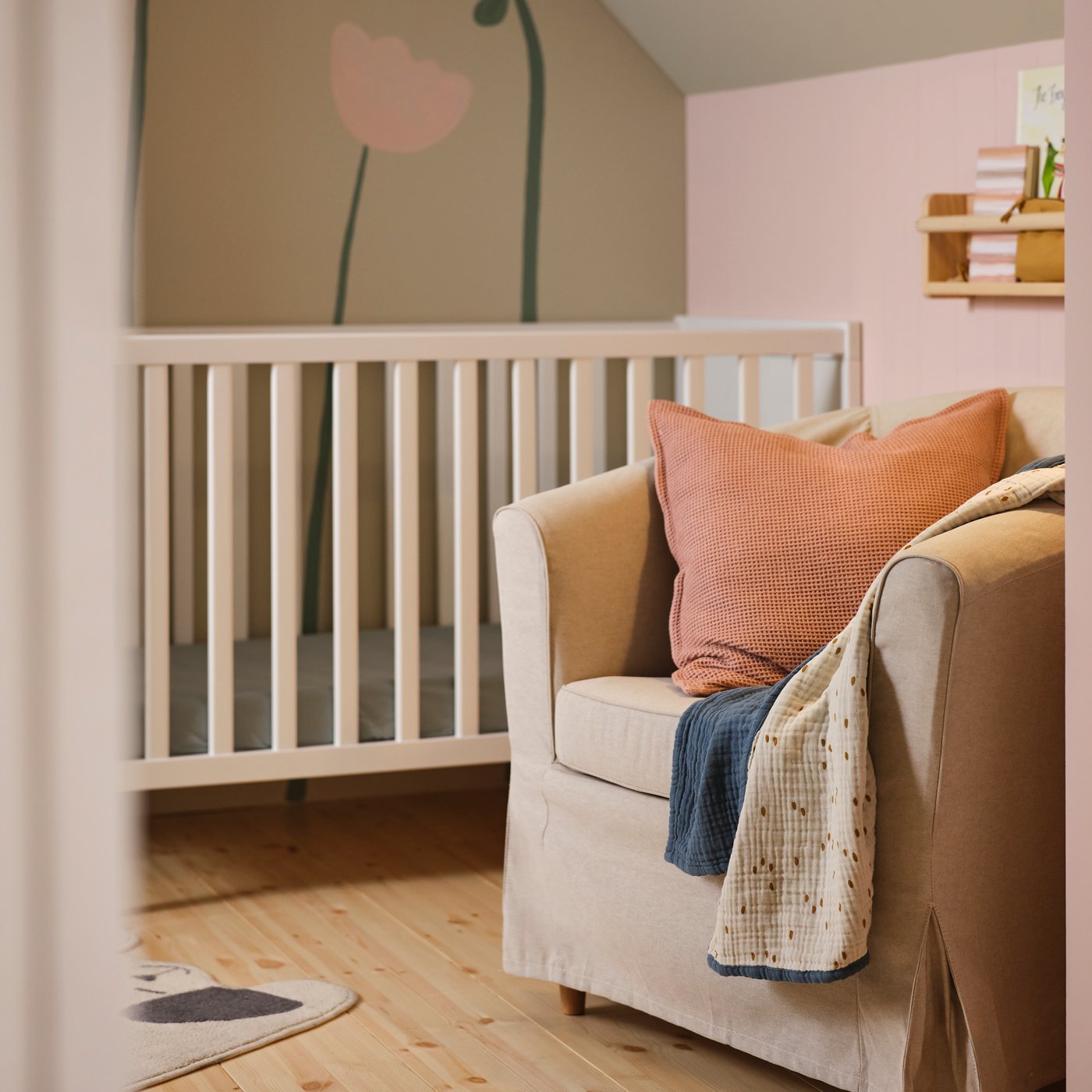 En TULLSTA fåtölj med beige LOFALLET överdrag, en GULDVÄVARE pläd och en kudde med rosa överdrag, placerad intill en SUNDVIK spjälsäng.
