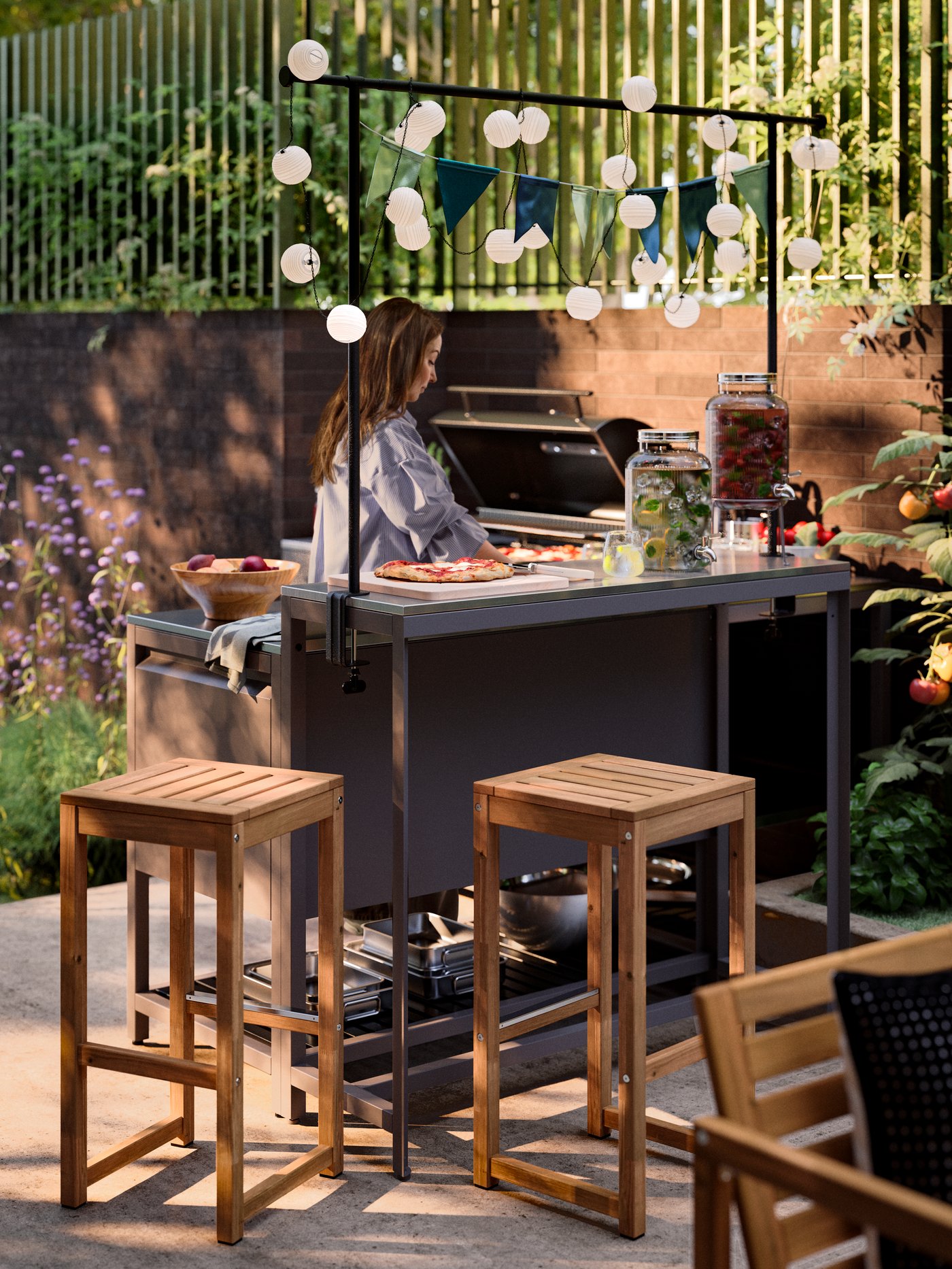 Two light brown stained NÄMMARÖ outdoor bar stools next to a BÅTSKÄR outdoor kitchen and bar table with a decorating rod.