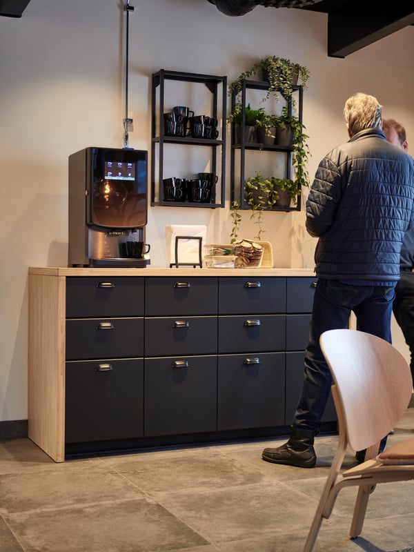 Kaffehörna i offentlig miljö med modern inredning, kaffemaskin, muggar, växter och två personer i samtal.
