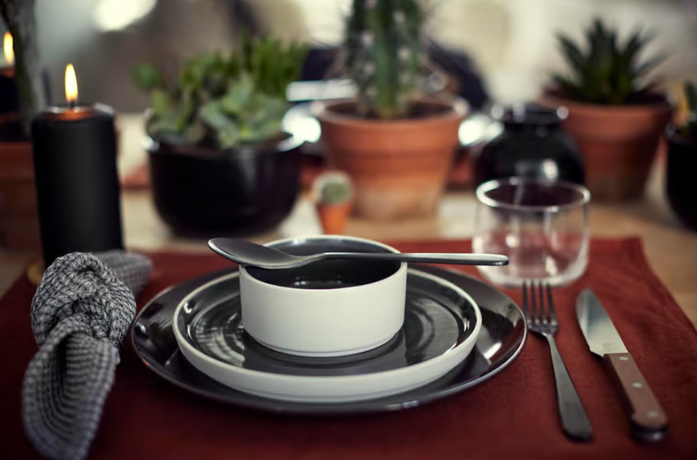 Gedeckter Tisch mit schwarzem Geschirr, rotem Platzset, Glas und Besteck, umgeben von Sukkulenten und einer brennenden Kerze.