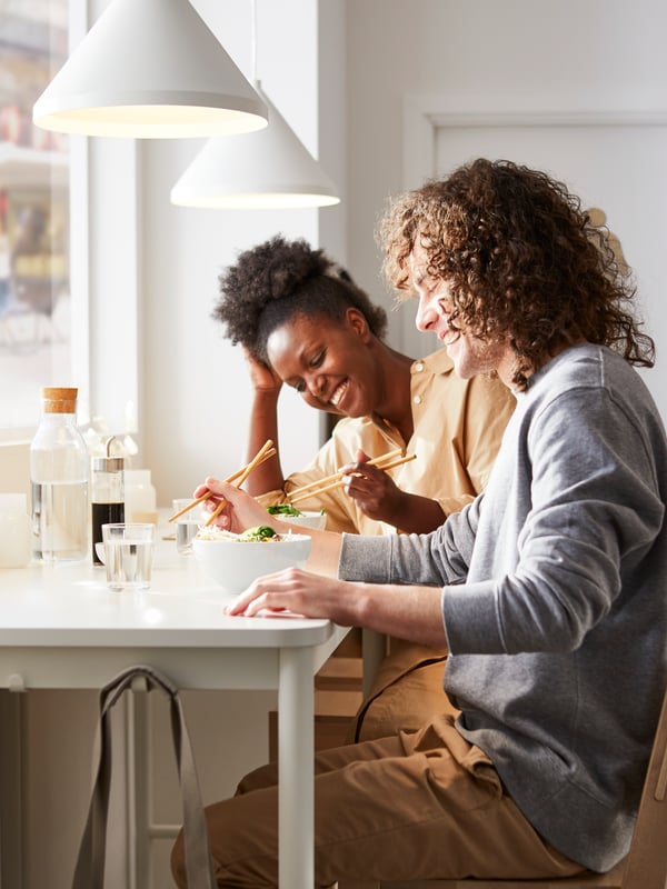 Two people having lunch together, smiling and holding chopsticks over two bowls of food on a TOMMARYD table by a window.