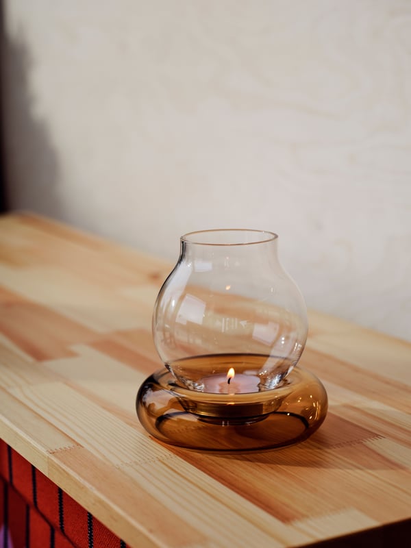 An OMMJÄNGE tealight holder in clear glass and brown-tinted glass sits on a bright wooden surface by a wall.