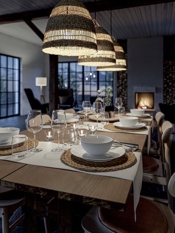 A wooden table set with white bowls and plates, glasses, cutlery, woven place mats and tealights.
