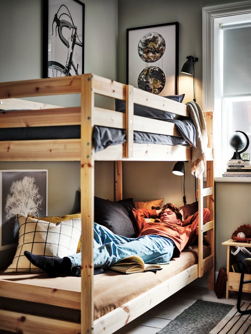 Children's room featuring a beige bunk bed frame with a person laying in the bottom bunk, cushions, wall frames, and a stool.