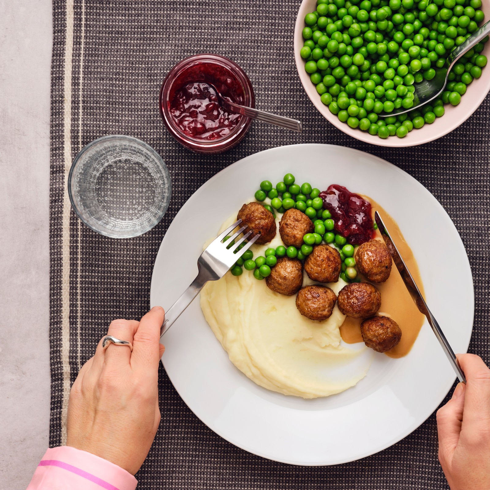 Osoba trzymająca widelec i nóż zabiera się do porcji klopsików, puree ziemniaczanego i zielonego groszku na białym talerzu.