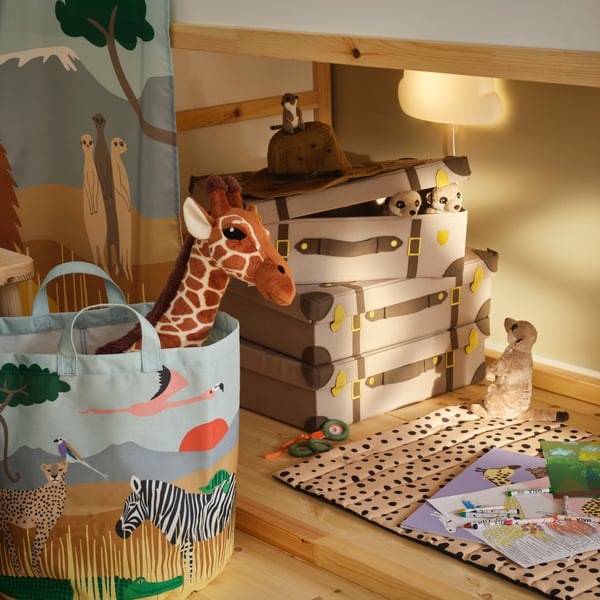 Three storage trunks are stacked under a KURA bed and a quilted mat lies on the floor with a SANDLÖPARE activity book on it.