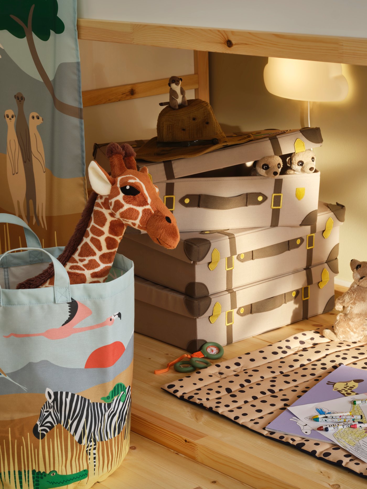 Three storage trunks are stacked under a KURA bed and a quilted mat lies on the floor with a SANDLÖPARE activity book on it.