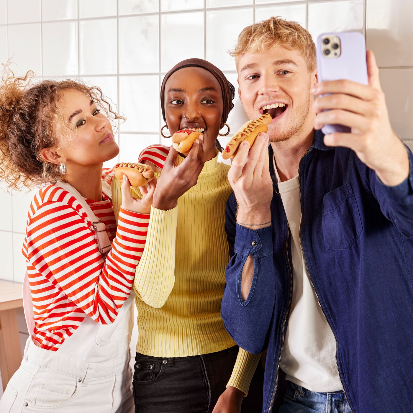 En gruppe unge spiser en hotdog mens de tager en selfie foran en væg med hvide fliser.
