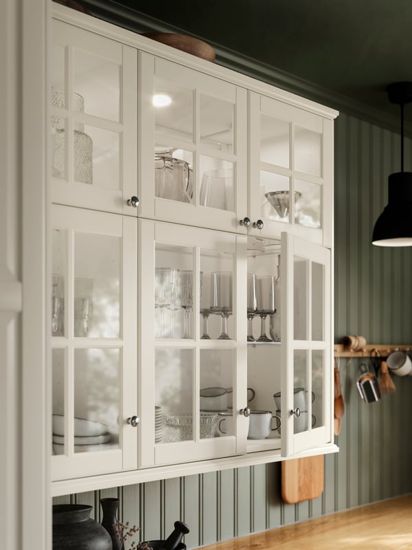 Una cucina con mobili con ante in vetro BODBYN bianco sporco e scaffali UTRUSTA su cui sono appoggiati tazze, bicchieri e articoli da cucina - IKEA.