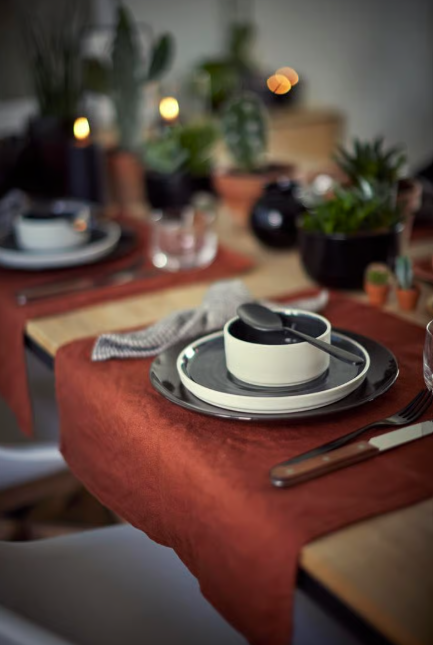 Gedeckter Tisch mit schwarzem Geschirr auf rotem Tischtuch, begleitet von Besteck, Glas und Pflanzen im Hintergrund.