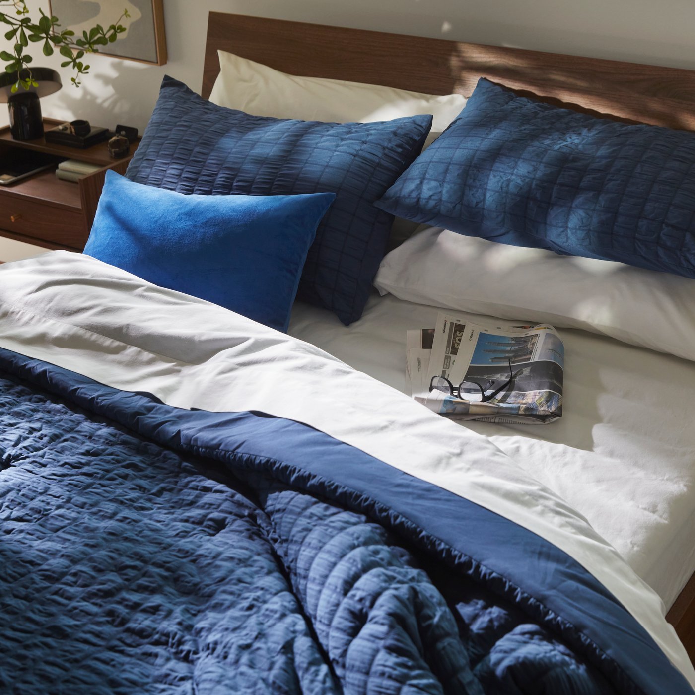 A dark blue RÄVSTARR comforter set with matching pillows is neatly laid on a bed, with a newspaper and glasses resting on it.