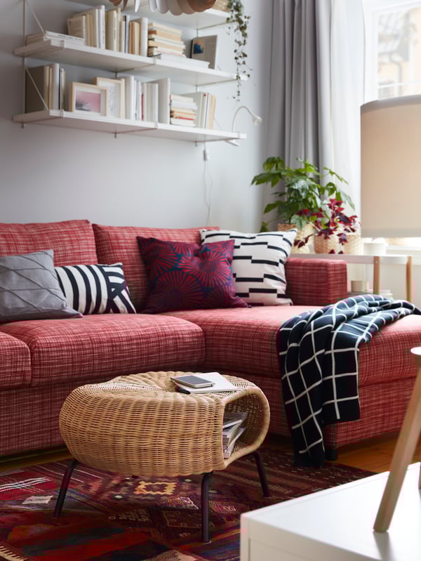 GAMLEHULT rattan footstool with storage on a red patterned rug, a red sofa, white wall-mounted shelves and a checkered throw.