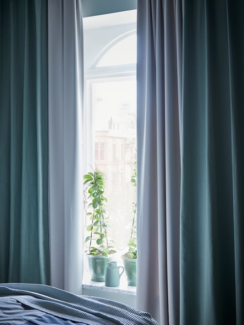 A bedroom window with MAJGUL room darkening curtains in light grey under blue curtains open to show plants in the windowsill.