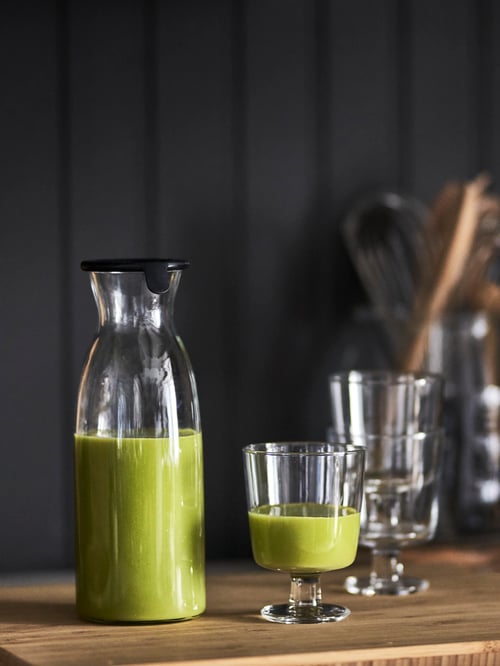 View of kitchen countertop, glass goblet, carafe with lid, containing green smoothie, stacked goblets and cooking utensils.