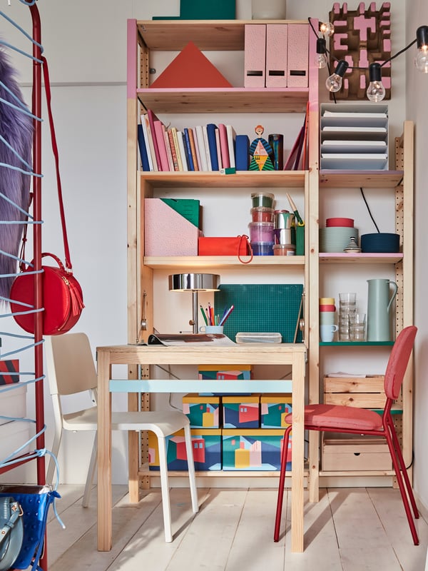 An open shelving unit holding various books, files, baskets, and dinnerware, with a table and two chairs in front.