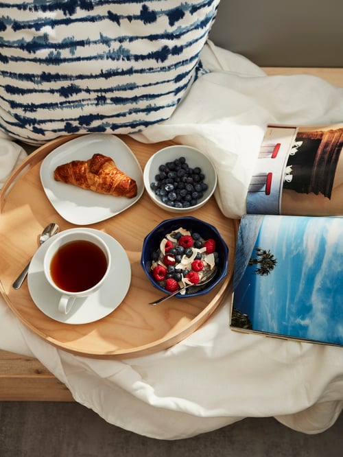 SKALA Tablett auf einem Bett, darauf eine VÄRDERA Teetasse mit Untertasse und einer STRIMMIG Schüssel mit Müsli.