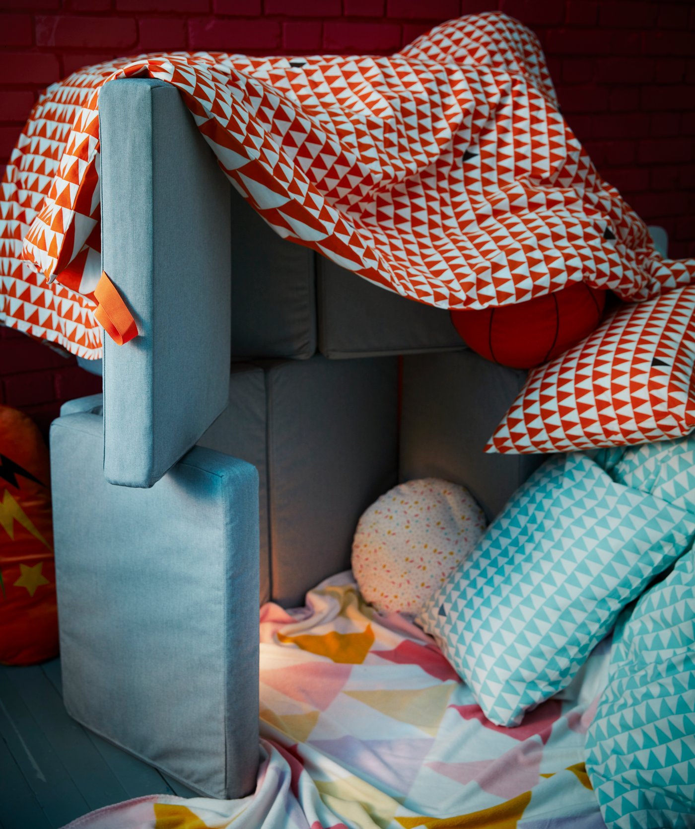 A hut or fort built with grey SLÄKT cushions, a red duvet cover, and blue pillows.