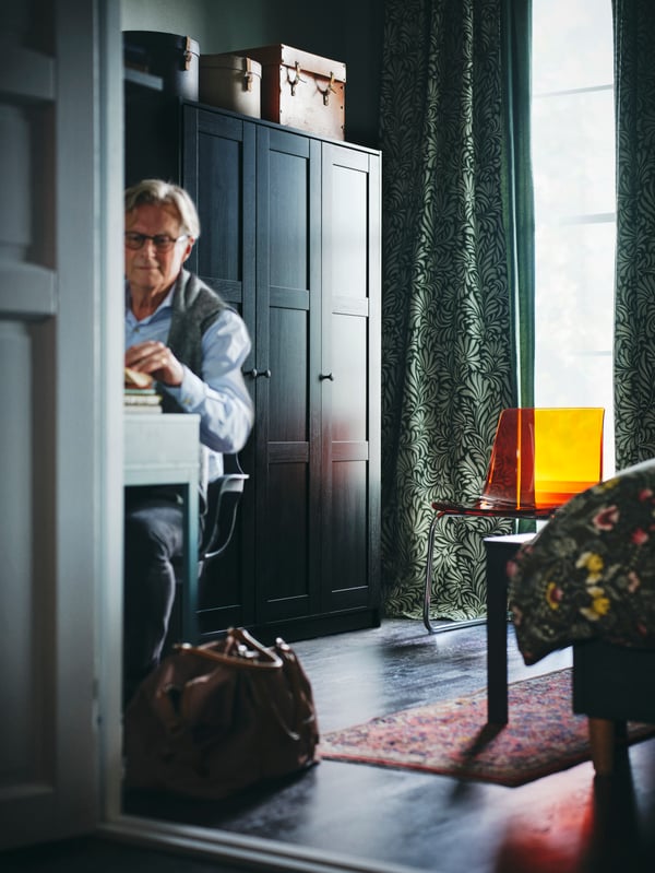 A person in a room with a black-brown RAKKESTAD wardrobe with three doors and dark green/leaves TRÅGSPINNARE curtains.